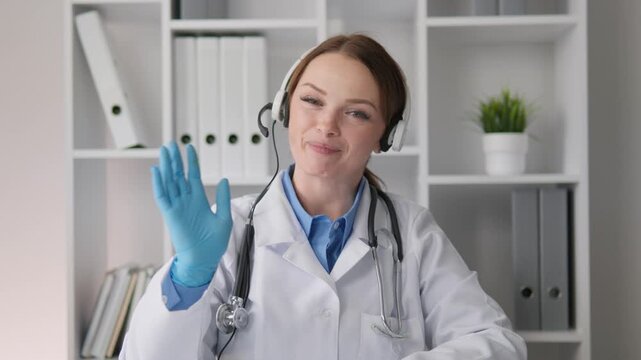 Young woman doctor wearing gloves and headset communicates with patient via video call in bright medical office. Telehealth and remote healthcare services concept for modern medicine