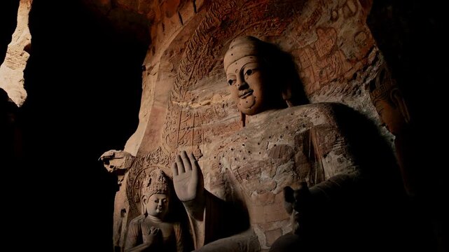 View of the ancient Yungang Grottoes Buddha statue with intricate carvings and serene expression inside a dark rock cave in Datong, Shanxi, China.