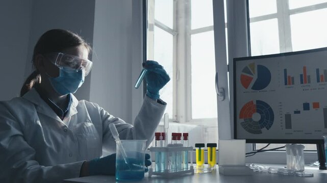 Female researcher conducts chemical analysis using test tubes while monitoring scientific data on computer in bright laboratory. Innovation, safety, data driven science are highlighted in this
