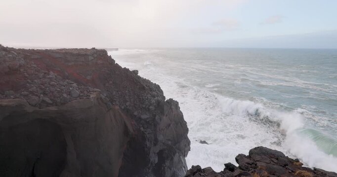 View of the rugged volcanic cliffs of Snaefellsnes with powerful white waves crashing against the dark rocks under an overcast sky in Eyja- og Miklaholtshr, Iceland.