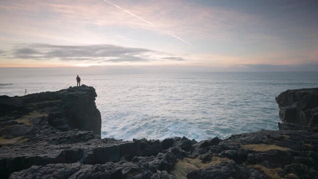 View of a photographer standing on the rugged rocky cliffs of Snaefellsnes overlooking the ocean waves during a soft sunset in Eyja- og Miklaholtshr, Iceland.