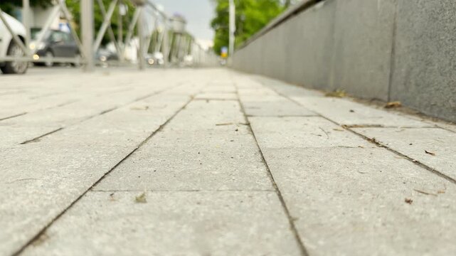 Cars scurry along the city road behind a metal fence. A paved sidewalk stretches forward, guiding the view toward the busy urban background.