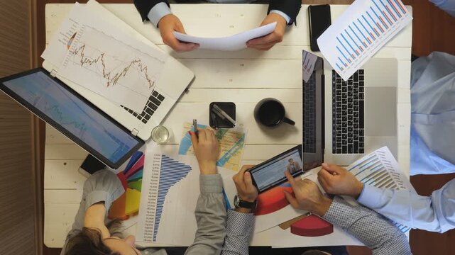 Young tired coworkers analyzing financial graphs at office. Group of exhausted colleagues work on business development sitting at table. Top view to male and female hands of managers. Slow motion