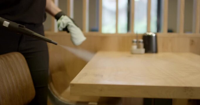 Worker using high pressure steam nozzle to disinfect wooden restaurant table surface with vapor mist