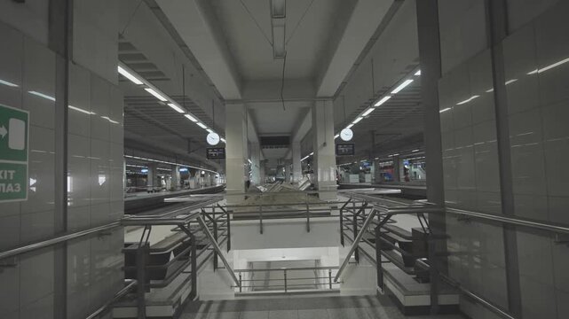Camera zooming out from stairs at modern underground train station platform with digital clocks and directional signs.