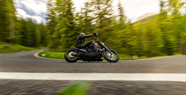 Custom motorcycle leaning through a winding mountain road surrounded by forest. Dynamic panning image with motion blur highlights speed, freedom and motorcycle touring adventure.