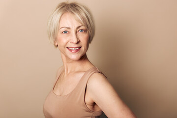 Fototapeta premium Smiling mature woman with short blonde hair wears beige tank top against neutral background. Studio portrait with soft lighting captures confident and natural beauty concept for aging gracefully.