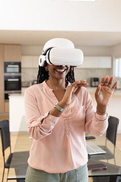 Adult woman wearing white VR headset and pink blouse, gesturing in kitchen with laptop