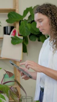 Vertical video: Holding tablet, caretaker in white top checking plant care in studio with plants