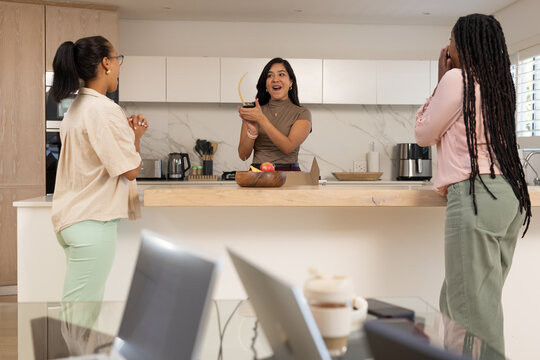 Diverse female friends celebrating around kitchen island, Indian woman holding small curved trophy