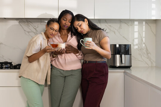 Diverse female friends leaning together, holding mugs and glass teacup in kitchen near cooktop