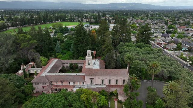 Historic Spanish mission church with bell tower and red-tiled roofs nestles among towering trees and lush green lawn under overcast skies in Santa Clara, California, USA. Aerial drone wide shot