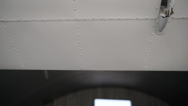 Close up video of an aircraft wing underside showing rivets and structure. Camera slowly zooms in on the metal skin and fasteners. Background shows hangar interior with soft lighting