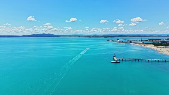 A boat moves across the clear water, leaving a wake behind it. A pier extends into the water, with land and hills visible far away. The sky is bright with few clouds.
