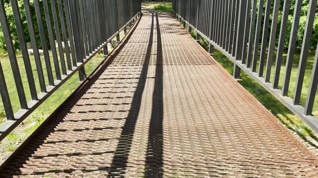 A metal pedestrian bridge extends over a river in a park. The grated walkway features long, parallel shadows cast by the railing. The scene is bright and sunny.