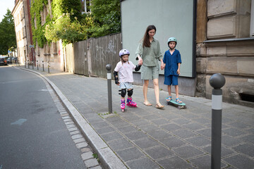 Fototapeta premium Mother supervises children rollerblading and skateboarding on a sunny city sidewalk