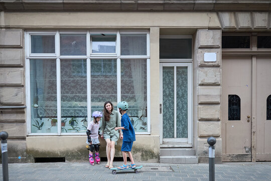 Mother watches her children enjoy outdoor activities on a city sidewalk