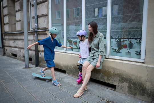 Mother watches children skateboarding and rollerblading on a city sidewalk