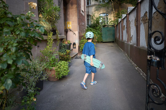 Young boy in helmet holding a skateboard walks down a narrow alleyway with plants