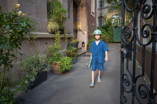 Young boy in blue outfit with skateboard walks through a gate in a courtyard