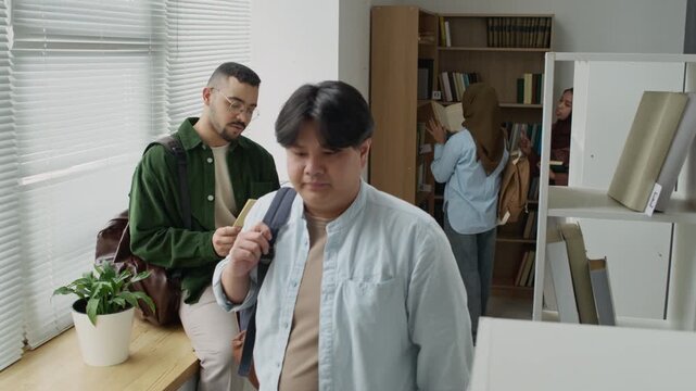Middle Eastern young adult male student reading book in library with Asian classmate walking past and Muslim female student browsing bookshelf. Clip perfect for marketing diverse higher education