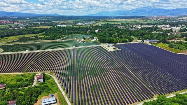 A large solar panel field stretches across the land near a small town. The panels are arranged in rows and reflect the sun. Mountains rise in the background under a blue sky.