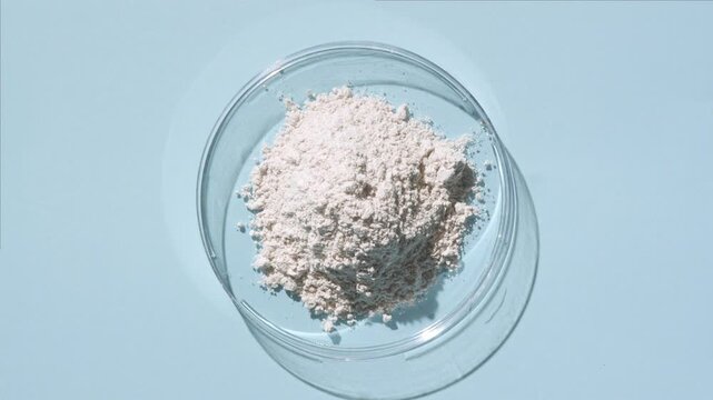 View from above capture of a pile of white fine powder in a plastic petri dish on a pale blue background lit by moving directional light casting dynamic shadows