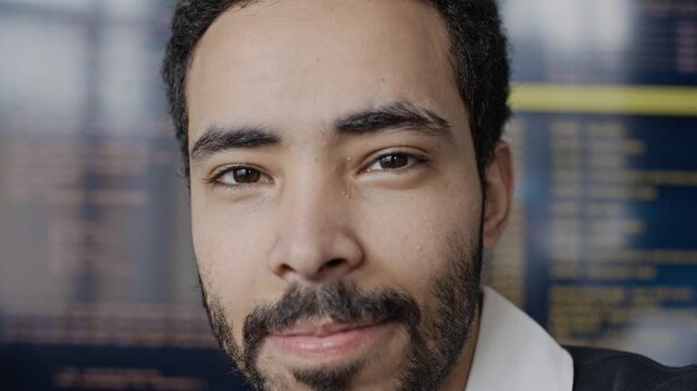 Closeup slowmo portrait of young Middle Eastern male programmer looking at camera with computer code on screen in background. For IT recruitment, software development ads, and tech company branding