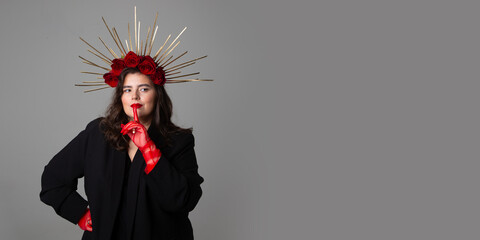 Obraz premium Photo banner. Young adult white woman with make up, black jacket, DIY golden halo crown, rose on head, red tulle gloves, choker, collar on neck raised index finger to lips on gray. Beauty. Copy space