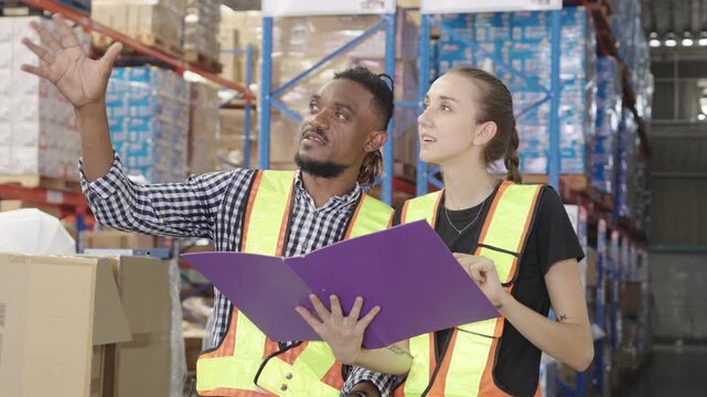 Two employees meticulously review purple folder within brightly lit warehouse environment surrounded by numerous stacked cardboard boxes; wearing safety vests and casual clothing, they conduct quality