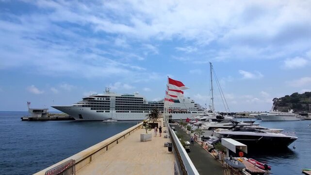 Monaco, Monaco - 21 June 2026: View of a large cruise ship docked at the harbor with luxury yachts and the coastline visible under a blue sky.