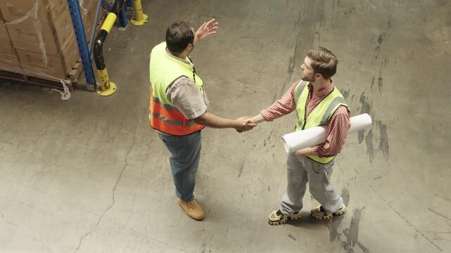 Two construction workers collaborate on warehouse plans discussing logistics strategies within vast industrial storage facility featuring concrete flooring and racking systems.