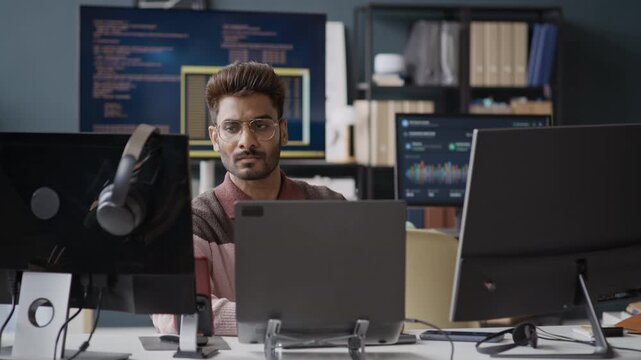Medium shot of Indian young man with glasses working on computers at desk in IT office, looking at screens and thinking. Perfect for software development, programming, and tech industry promotions