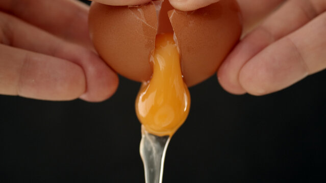 Close-up of hands cracking brown eggshell to release bright orange yolk into a metal spoon