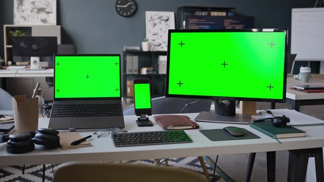 Pan shot of laptop, smartphone, and desktop monitor displaying green screens on modern office desk. Perfect for showcasing responsive web design or software mockups