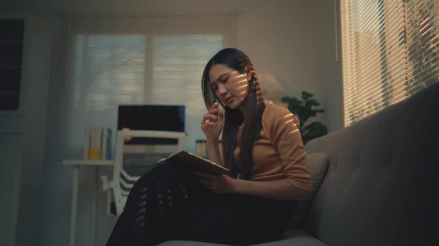 Frustrated Asian woman feeling tired and stressed while studying on the sofa, eventually lying down with the book covering her face in a dimly lit living room with sunlight from the window