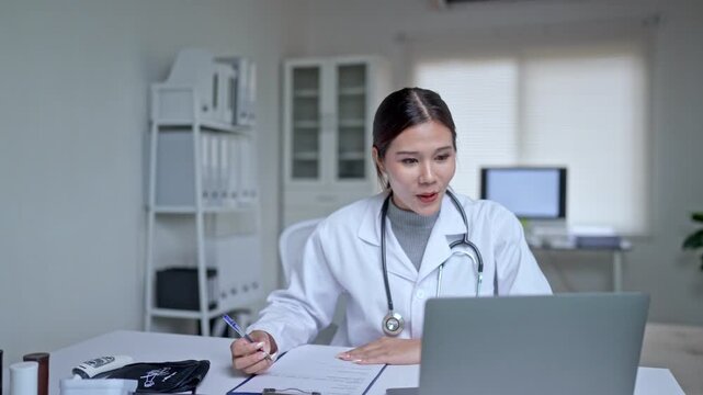 Professional asian female doctor in a white coat with a stethoscope providing an online consultation to a patient, talking on a video call on her laptop and taking notes in her modern office