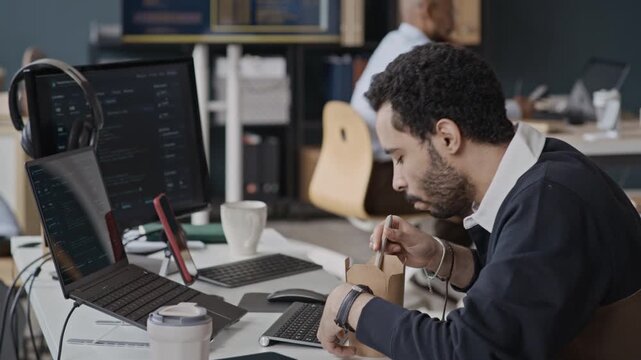 Side view handheld shot of young Middle Eastern man eating takeout noodles at office desk with computer screens showing code. Ideal for software developer lifestyle and busy workday concepts