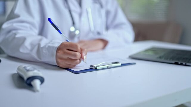 Close up of a female doctor in a white coat with a stethoscope writing a medical prescription or filling out a patient's health insurance form at her desk in a modern hospital clinic