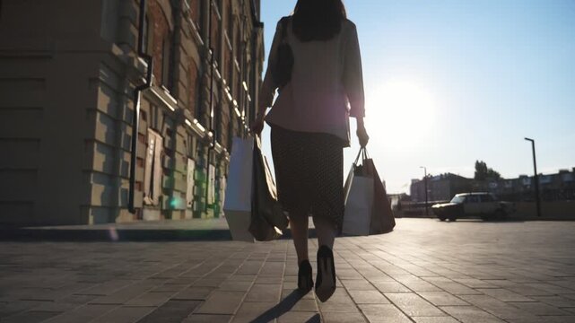 Young female customer holds shopping bags walking at sidewalk after purchases. Fashionable woman carries paper packets going along urban square. Concept of leisure and free time. Rear view Slow mo