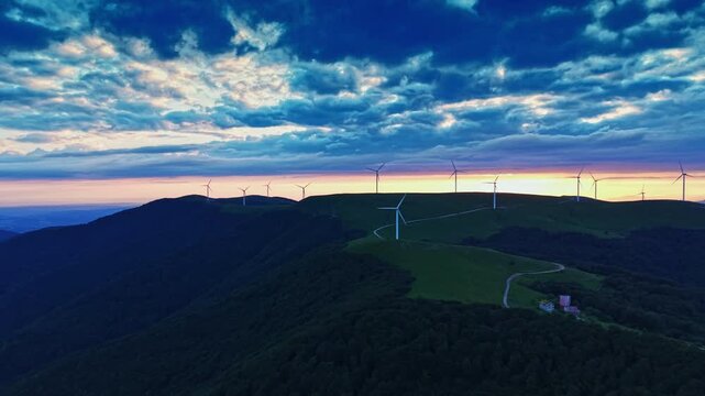 A landscape shows rolling hills with wind turbines lined up against a colorful sky at sunset. A small farm is located at the base of the hills. Clouds stretch across the sky.
