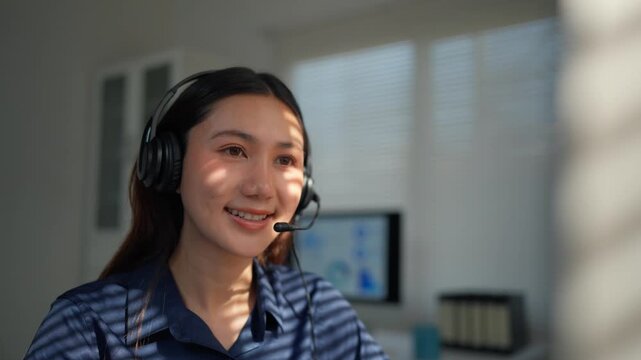 Friendly asian female customer service representative wearing a headset with a microphone, talking to a client in a sunlit office, providing helpful and efficient telemarketing assistance