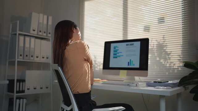 Asian office worker stretching her sore muscles after a long day of work, feeling tired and stiff from her sedentary job while sitting at her desk with a computer in front of her
