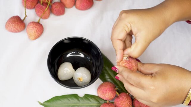 A woman hand Holding a Freshly Peeled Lychee Fruit, Revealing Its Juicy White Flesh and red Outer Shell on keep black bowl. inside with another whole lychee or litchi fruits.