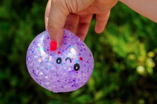 A kawaii dumpling shaped squishy toy with glitter texture and a cute cartoon face is squeezed gently by a finger with red painted nail outdoors