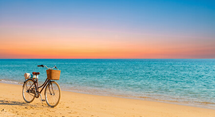 Fototapeta premium Vintage bicycle with a wicker basket parked on a sandy beach by a turquoise sea at sunset