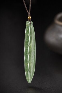 A slender green jade pendant shaped like a silique on a dark background.