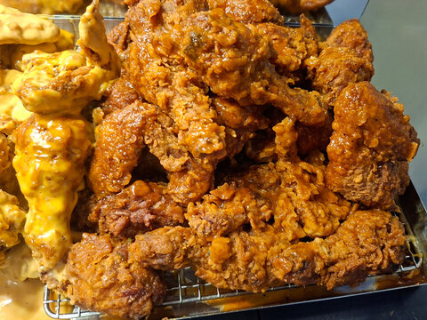 A heap of crispy fried chicken pieces drenched in a thick, glossy sweet and spicy sauce, alongside some pieces coated in a rich, melted yellow cheese sauce, arranged on a wire mesh tray.