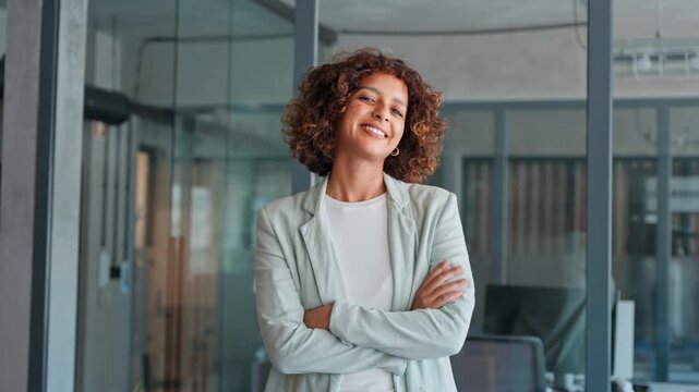 Portrait of smiling young multiracial latin or indian businesswoman with curly hair standing in office, wearing casual blazer in professional workspace. Arabian young woman girl smiling at camera