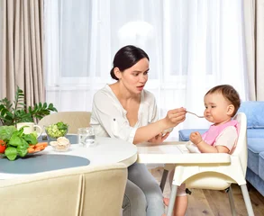 Mother feeding baby with a spoon. Spoon-feed. Happy young mother with a baby in the kitchen interior. Fresh vegetables and fruits. © Svetlana Fedoseeva